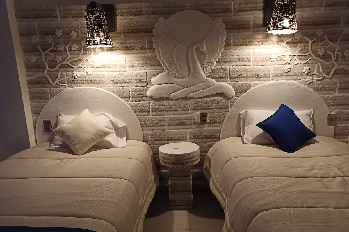 Een kamer met twee bedden, elk met hoofdbord en kussen. Daartussen staat een klein, rond tafeltje. De muur achter de bedden heeft een bakstenen patroon en decoratieve details.