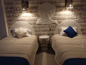Una habitación con dos camas, cada una con cabecero y almohada. Hay una mesita redonda entre las camas. La pared detrás de las camas presenta un diseño de ladrillo y detalles decorativos.