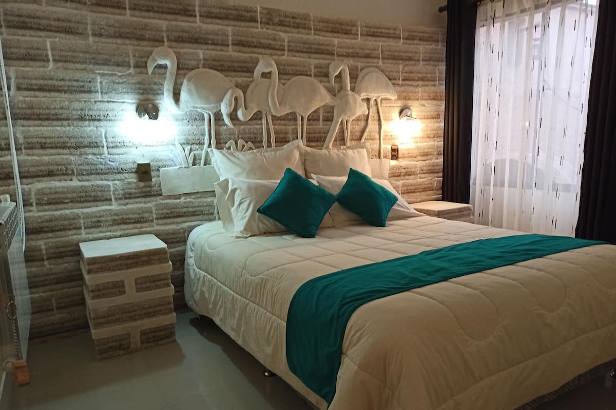Een slaapkamer met een hoofdbord met een baksteenmotief, een groot bed met wit beddengoed en turquoise kussens, een nachtkastje met een lamp, en een raam met dunne gordijnen.