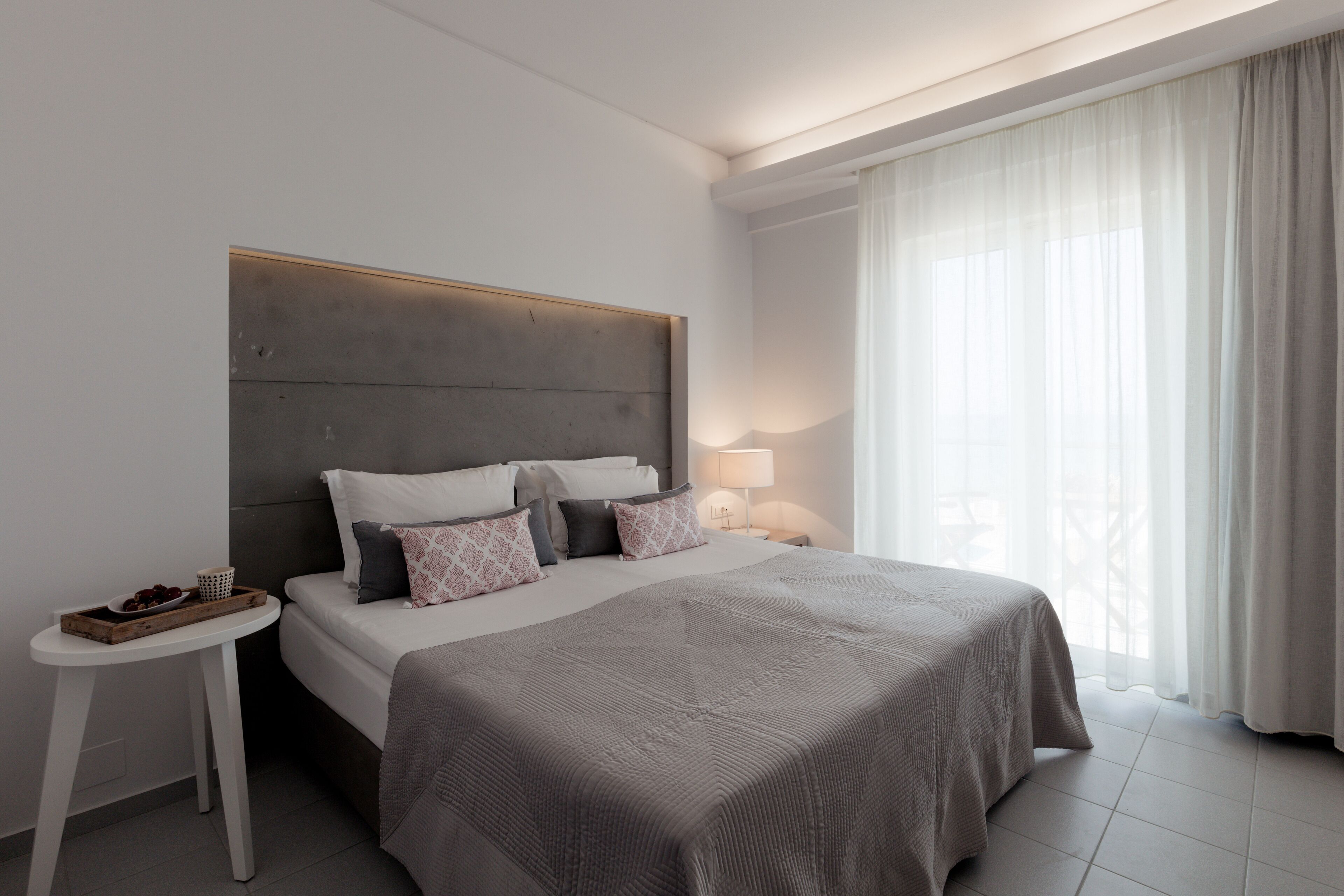 A modern hotel room with a large bed, a bedside table, and a view of the outdoors through sheer curtains.