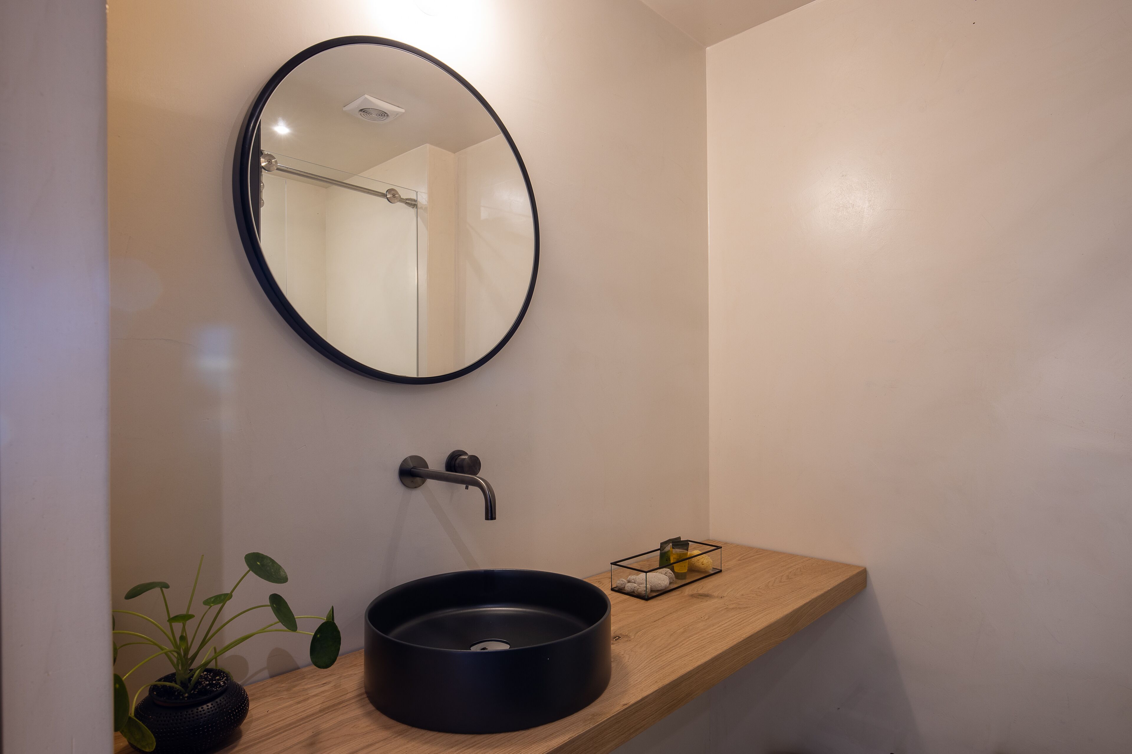 A modern bathroom with a round mirror, a black sink, a wooden countertop, and a potted plant.