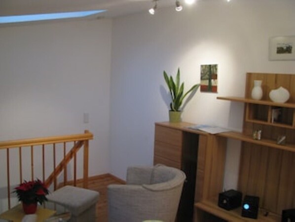 Ein Zimmer mit einer Holztreppe, einem grauen Sessel, einem kleinen Tisch mit Pflanze und einem Holzregal mit Dekorationen.