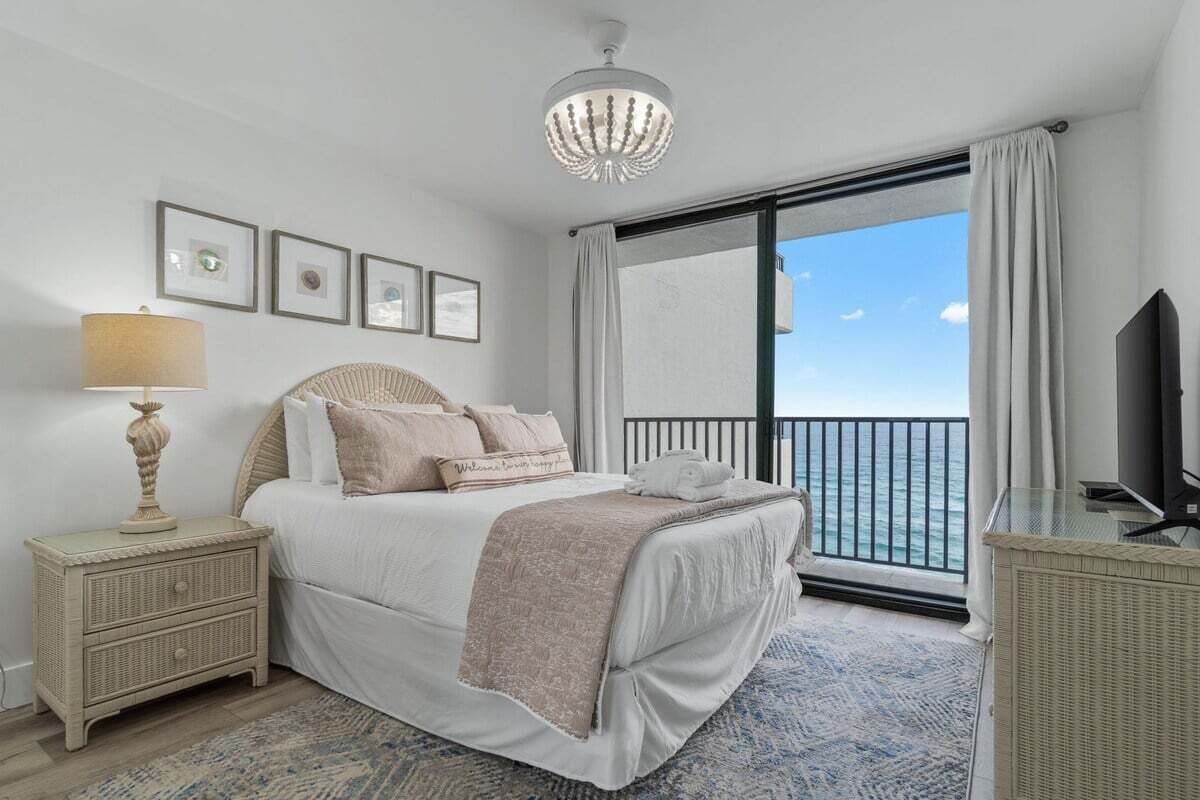 Un dormitorio con cama, mesita de noche, lámpara, televisor y balcón con vista al mar.