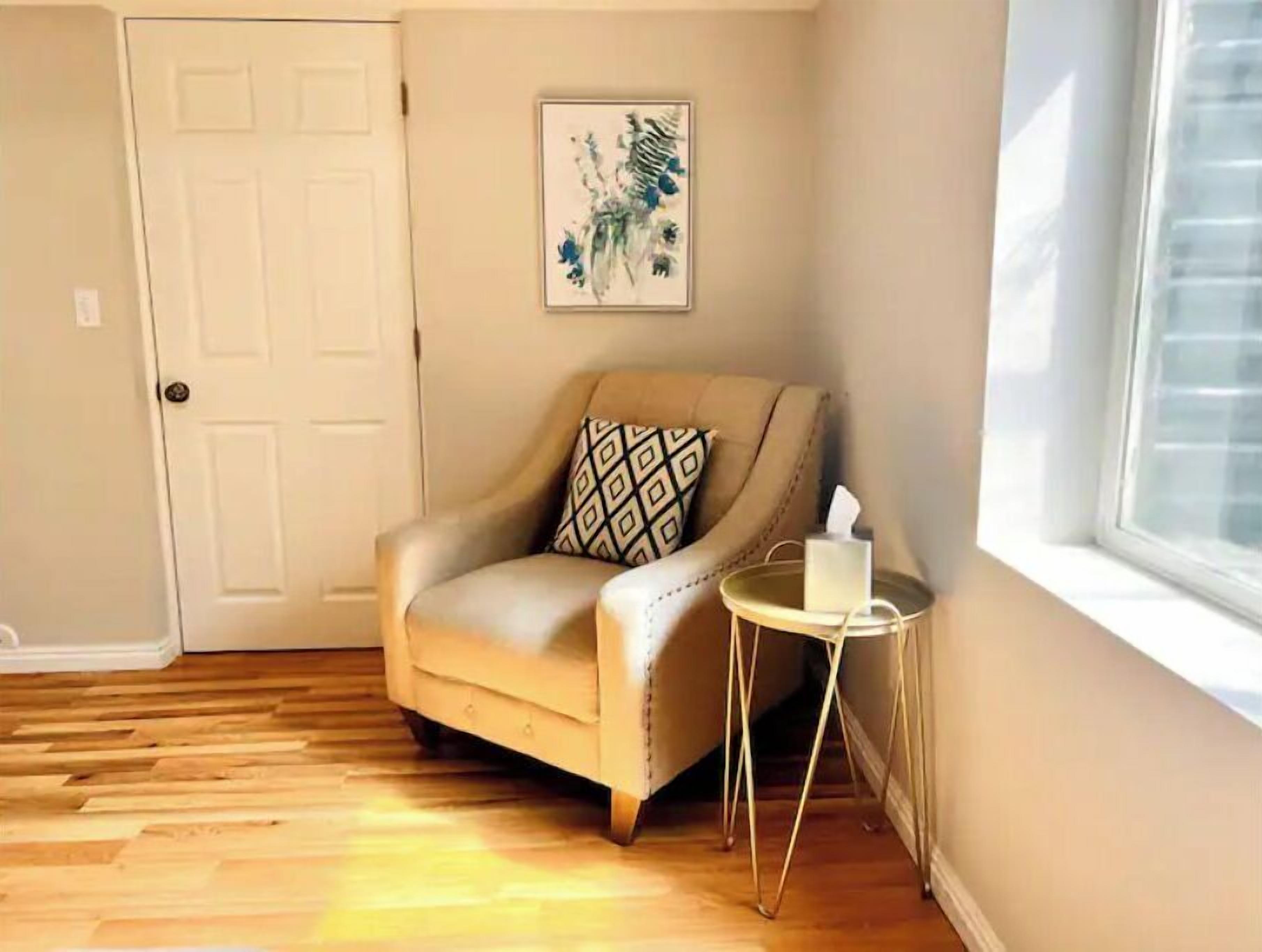 A corner room with a beige armchair, a small round table with a tissue box, a door, and a framed artwork on the wall.