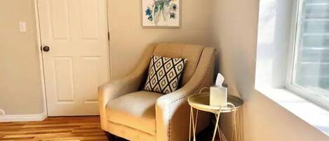 A corner room with a beige armchair, a small round table with a tissue box, a door, and a framed artwork on the wall.