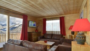 Una habitación acogedora con paredes de madera y un ventanal con vistas a la montaña. Hay un sofá, una mesa con un bol y una lámpara sobre un pedestal de madera.