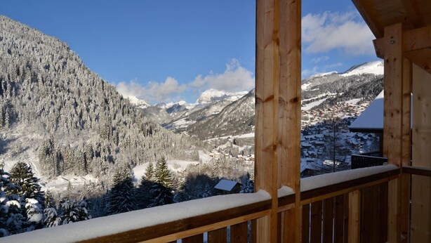 Un balcón de madera con vistas a montañas nevadas y un pueblo en la falda.