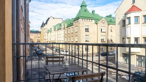 A balcony with wooden tables and benches, overlooking a street with parked cars and historic buildings with green roofs. - 1br apartment with balcony and sea view (Helsinki)