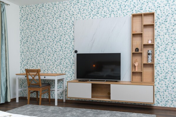 Une pièce comprenant un téléviseur posé sur un meuble en bois, un bureau avec une chaise et une étagère ornée d’objets décoratifs.