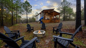 A cabin with a fire pit and Adirondack chairs in a wooded area.