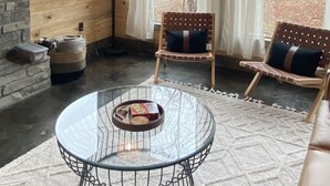 A cozy room with a stone fireplace, a glass coffee table, and a view of the forest through large windows.