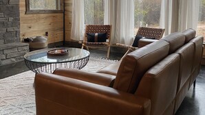 A cozy living room with a leather sofa, a glass coffee table, and a wicker chair. There are large windows with curtains, a stone fireplace, and a view of trees outside.