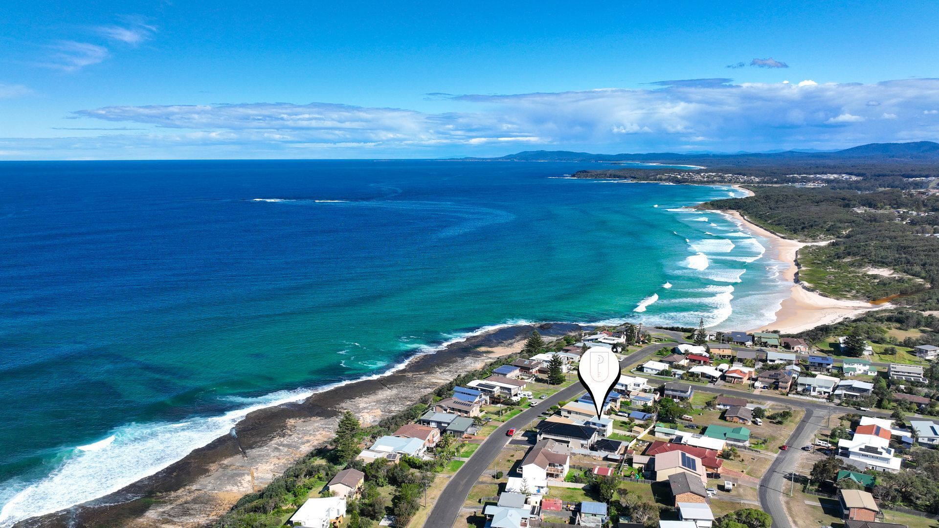 An aerial view of a coastal town with a beach, houses, and a clear blue sky.