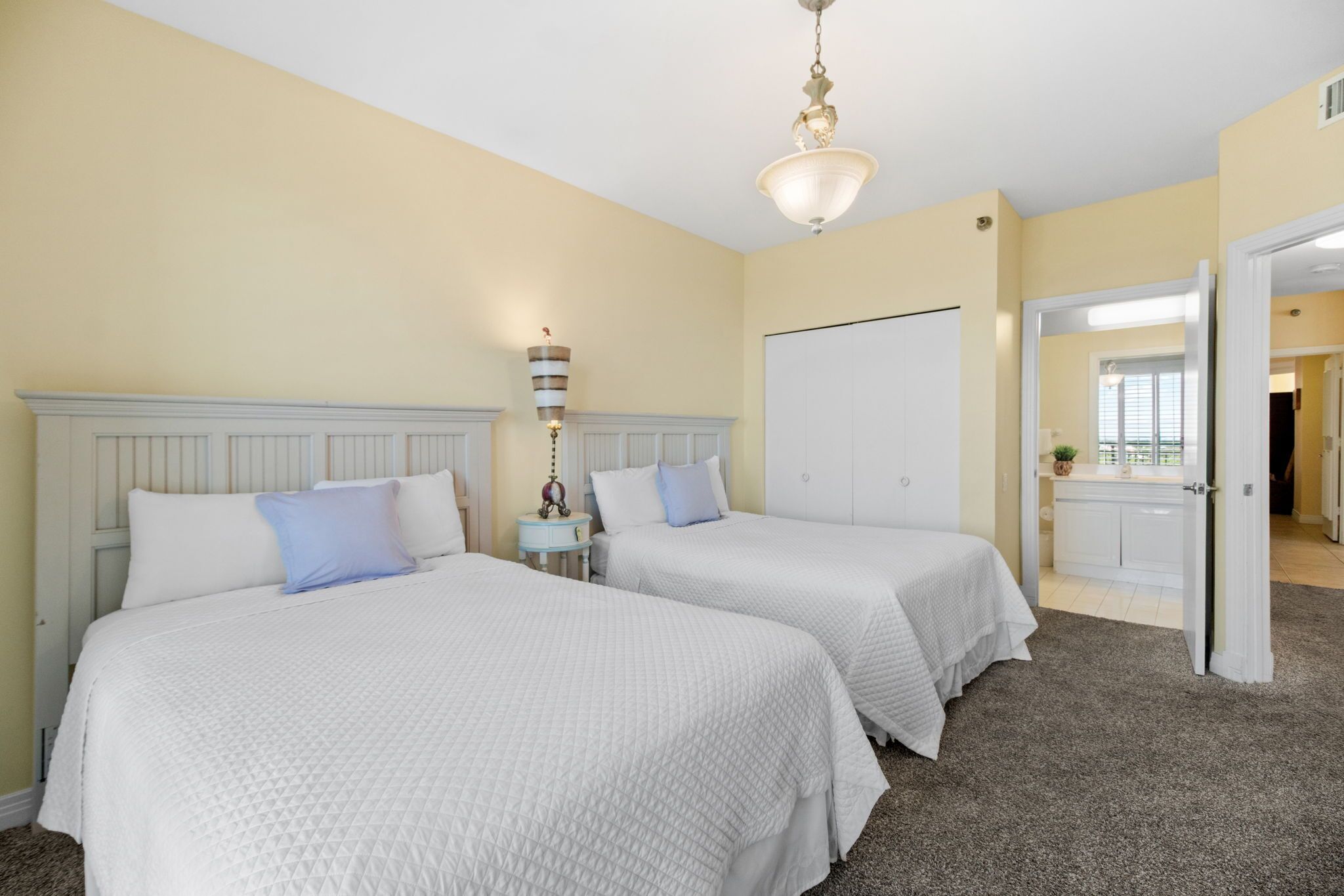 A hotel room with two beds, a white headboard, a bedside table with a lamp, and a view of the kitchen through an open door.