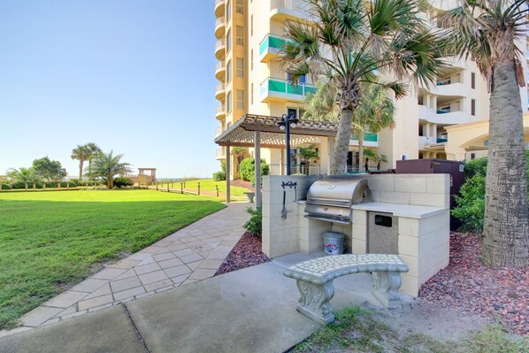 A patio area with a barbecue grill, a stone bench, and a pathway leading to a building with balconies and palm trees. - Beach Colony Tower 15A - Indoor Pool - HASSLE-FREE CHECK OUT (Perdido Key)