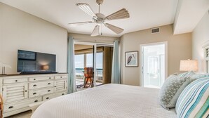 A bedroom with a large bed, a ceiling fan, a television on a dresser, and a view of the ocean through a sliding door.
