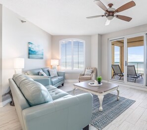 A spacious living room with a ceiling fan, a large window with blinds, a view of the ocean, and a balcony with outdoor furniture.