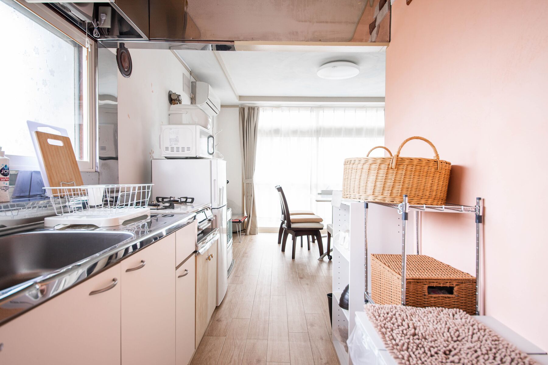 A compact kitchen with a sink, stove, and white cabinets. A chair and a wicker basket are visible in the background.