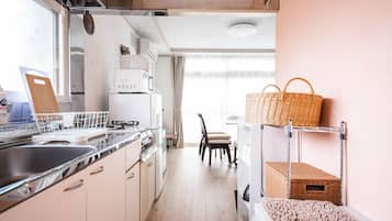 A compact kitchen with a sink, stove, and white cabinets. A chair and a wicker basket are visible in the background.
