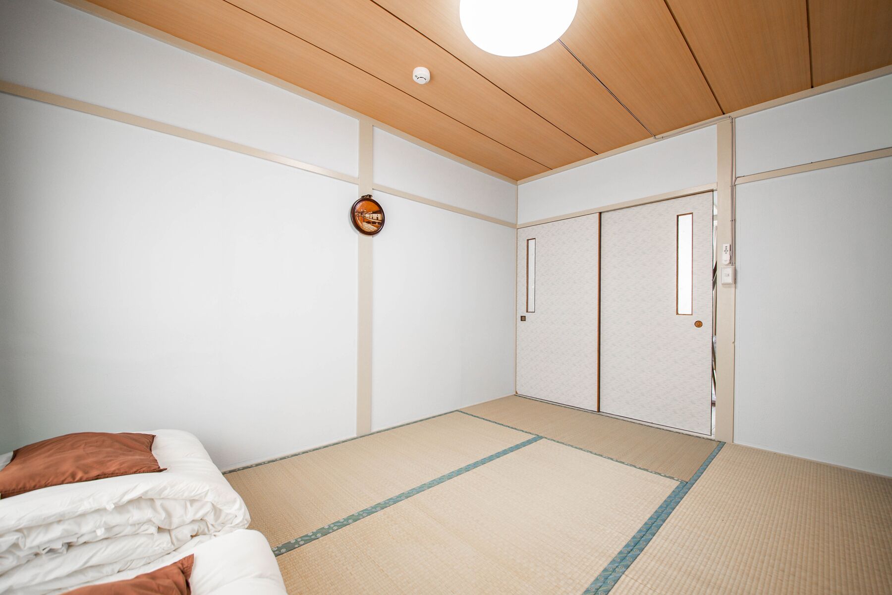 Une pièce avec un sol recouvert de tatamis, un lit avec une literie blanche et marron, un plafond en bois et une porte coulissante.