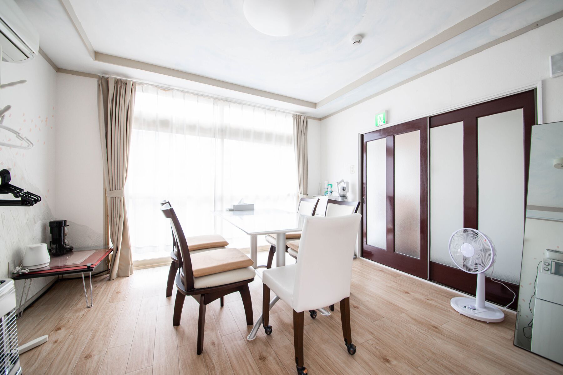 Ein Hotelzimmer mit Esstisch, zwei Stühlen, einem Standventilator und einem Kleiderschrank mit Spiegel.