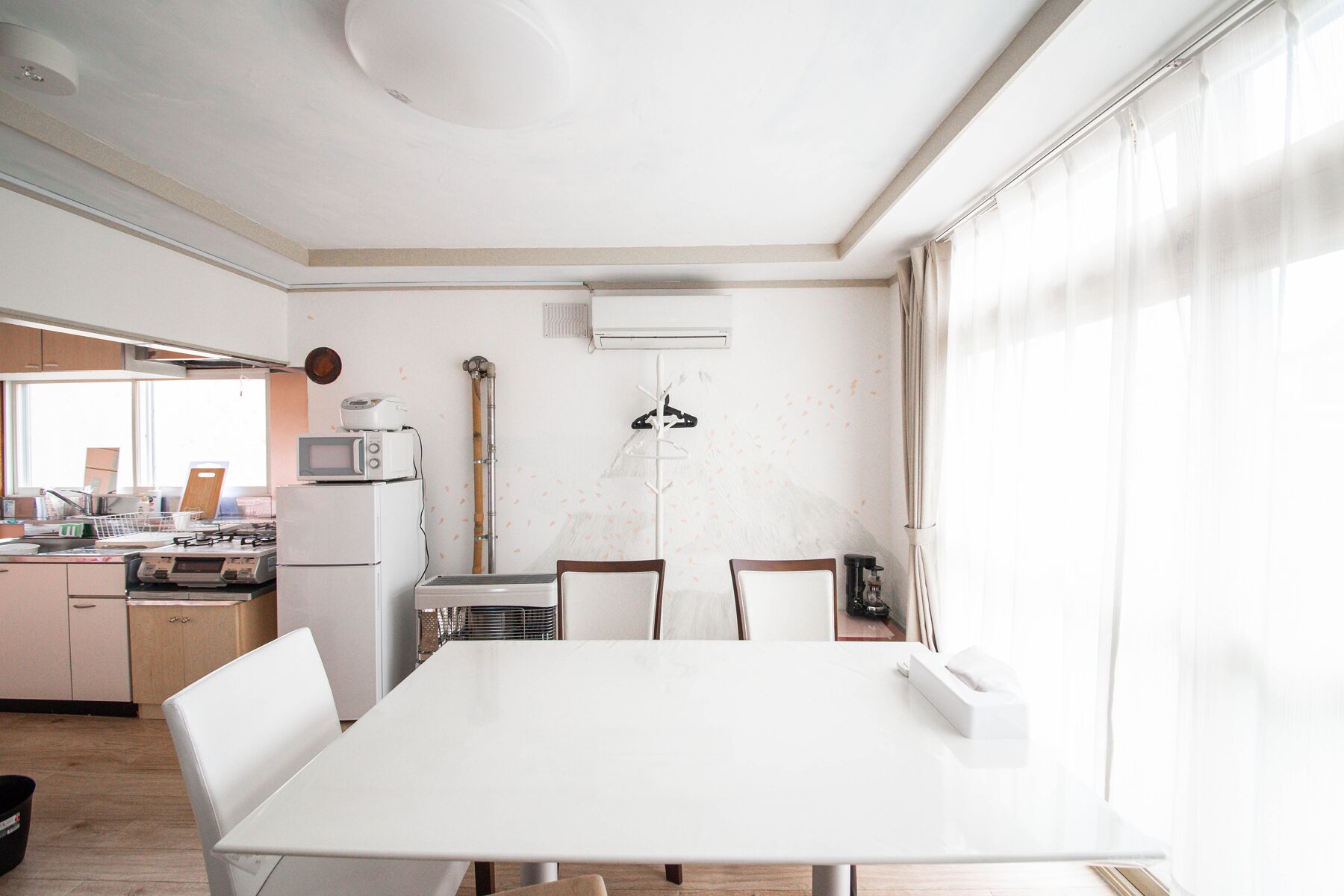A modern kitchen with white appliances, a dining table, and a large window with sheer curtains.