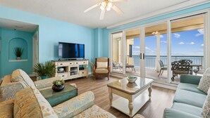 A spacious living room with a television, comfortable seating, a ceiling fan, and a balcony with a view of the ocean. - Beach Colony Tower 9C - Indoor Pool - HASSLE-FREE CHECK OUT (Perdido Key)