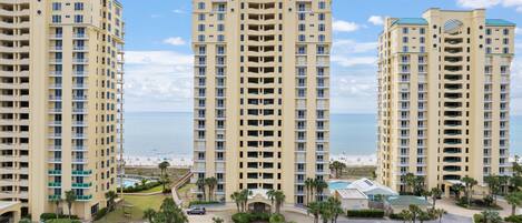 A high-rise hotel with multiple balconies, a parking lot, and a view of the beach.