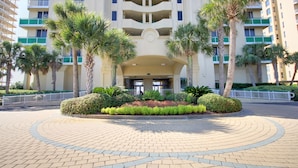 A hotel entrance with a circular driveway, palm trees, and a multi-story building with balconies. - Beach Colony West 13A - Indoor Pool - HASSLE-FREE CHECK OUT (Perdido Key)