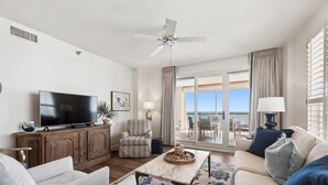 En rummelig stue med et fladskærms-tv, et træskab, en stribet lænestol, et sofabord og udsigt til en balkon med klar himmel.