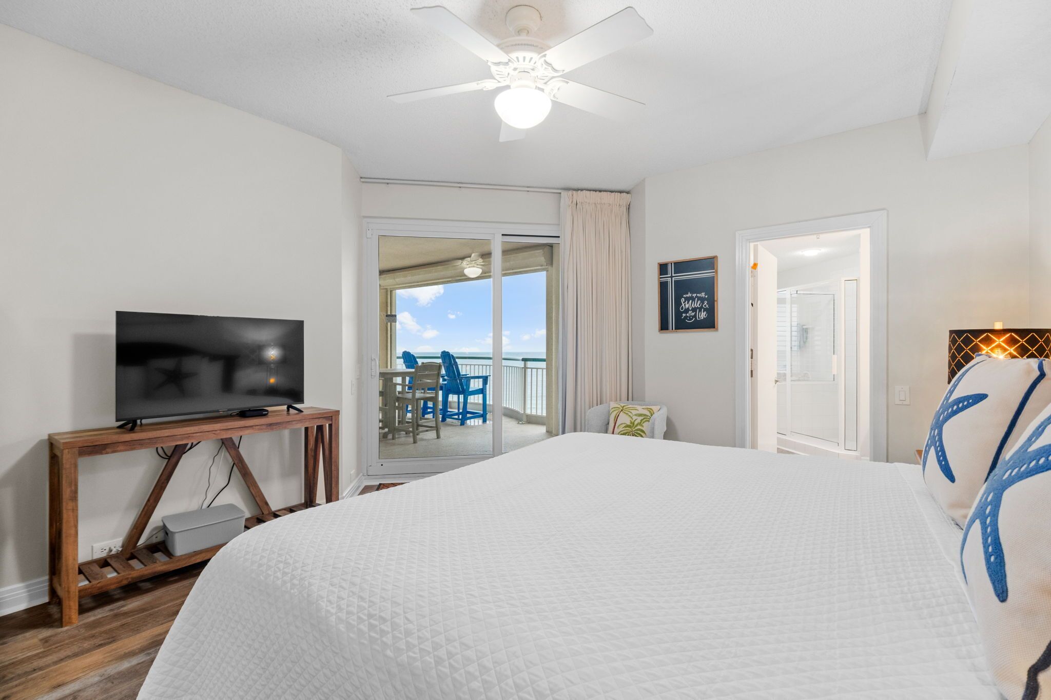 A hotel room with a large bed, a TV on a wooden console, a balcony with a view, and a ceiling fan.