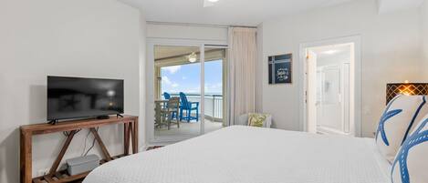 A hotel room with a large bed, a TV on a wooden console, a balcony with a view, and a ceiling fan.
