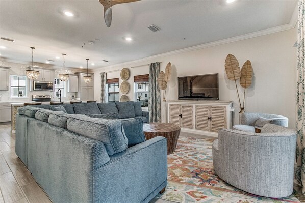 A spacious living room with a large sectional sofa, a round coffee table, and a grey armchair. A flat-screen TV is mounted on the wall, and there is a kitchen area in the background. - Lost Key Townhomes 14274 ~ Flip Flop Stop - NO HASSLE CHECK OUT (Pensacola)