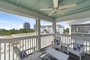 A covered outdoor area with white chairs and a ceiling fan, overlooking a road and buildings.