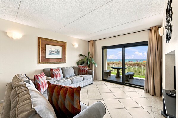 A living room with a sofa, a television, and a view of the beach through a glass door.
