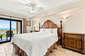 A hotel room with a large bed, a wooden headboard, a nightstand, a dresser, and a view of the beach through a sliding door.