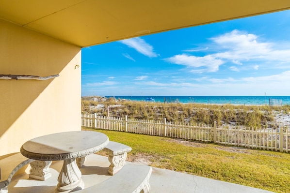 Een terras met een tafel en stoelen, met uitzicht op een strand met een witte hek en een helderblauwe lucht.
