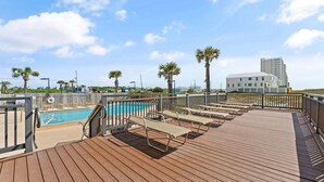 Um deck de madeira com espreguiçadeiras e piscina, cercado por palmeiras e um edifício moderno ao fundo.