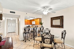 A dining area with a round table and chairs, a ceiling fan, and a kitchen in the background. - Ocean Breeze West Ground Floor -Beachfront- NO HASSLE CHECK OUT (Pensacola)