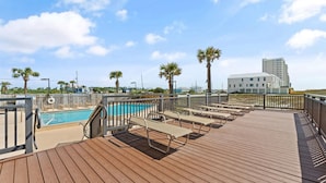 A wooden deck with lounge chairs and a pool, surrounded by palm trees and a modern building in the background.