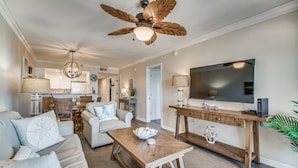 Una sala de estar amplia con ventilador de techo, televisor, mesa de centro de madera y un sofá con cojines decorativos.
