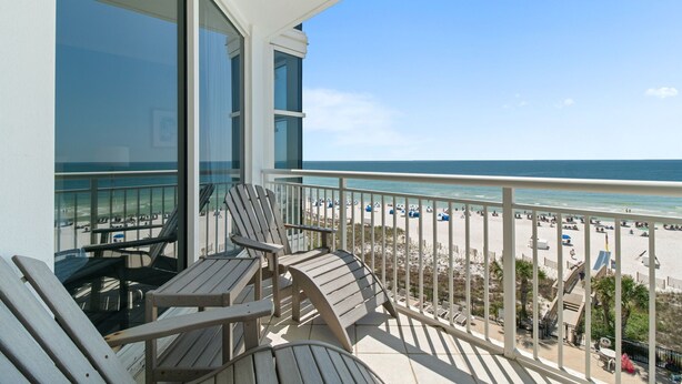A balcony with wooden chairs and a table, overlooking a beach with umbrellas and people.