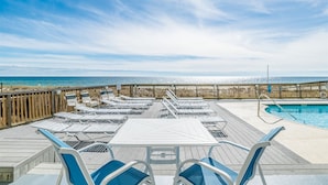 Een zwembadgedeelte met ligbedden en tafels, een houten terras en uitzicht op zee.