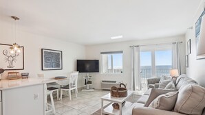 Een moderne woonkamer met een witte bank, een eethoek met een witte tafel en stoelen, een keuken met een wit aanrecht en een groot raam met uitzicht op zee.
