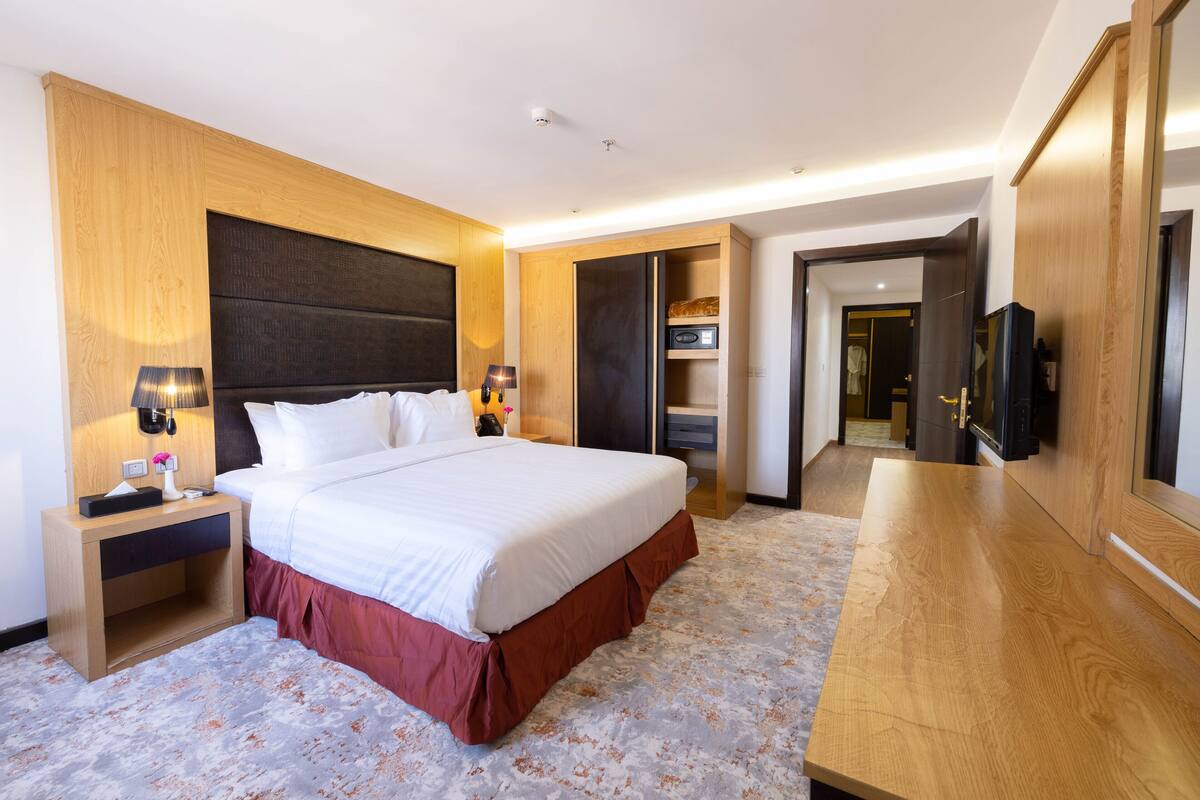 A modern hotel room with a large bed, wooden headboard, bedside tables with lamps, a built-in TV, and a wardrobe.