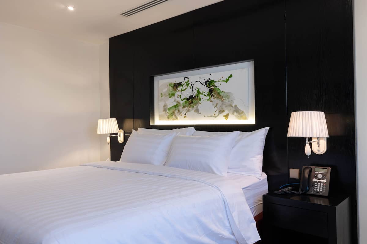 A neatly made bed with white linens, a bedside table with a lamp, and a wall-mounted phone.