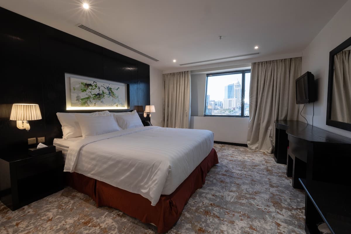 A hotel room with a large bed, two bedside tables with lamps, a flat-screen TV, a window with curtains, and a cityscape view.