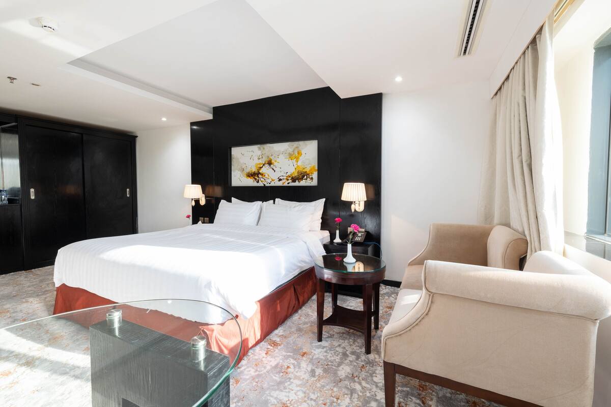 A hotel room with a large bed, two bedside tables with lamps, a glass coffee table, a beige armchair, and a black headboard with a painting.