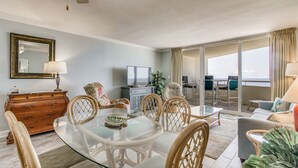 A spacious living area with a glass-top dining table surrounded by wicker chairs, a wooden cabinet, and a television. - Perdido Sun 1004 - indoor pool - Beachfront - NO HASSLE CHECK OUT (Perdido Key)