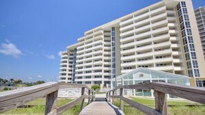 A multi-story hotel with a central glass elevator shaft, a wooden boardwalk leading to the entrance, and a clear blue sky. - Perdido Sun 1004 - indoor pool - Beachfront - NO HASSLE CHECK OUT (Perdido Key)